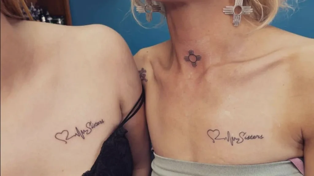 Two women featuring matching fine-line Memorial Handwriting Tattoos on their upper chests with a "Sisters" script and heartbeat design.