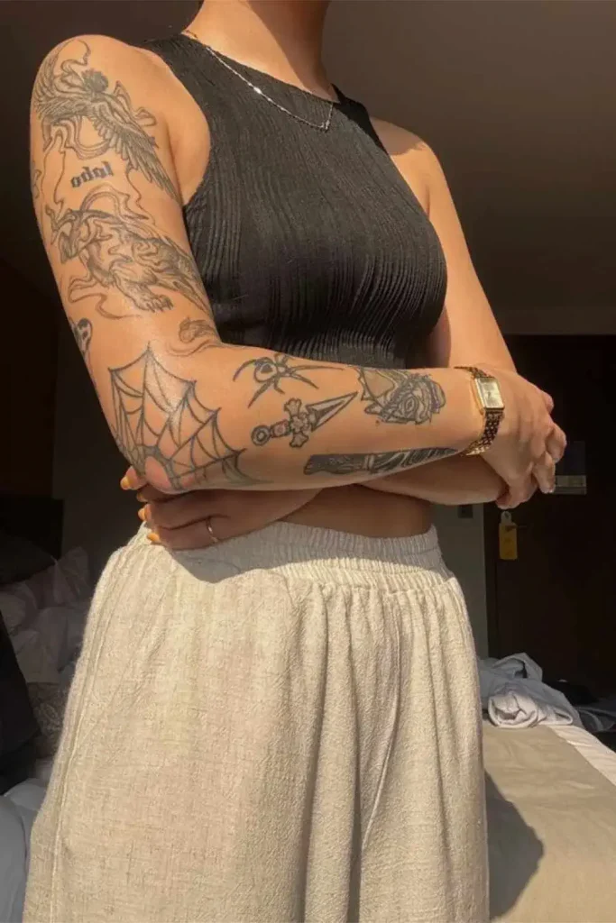 A woman modeling cohesive Y2K sticker sleeve tattoos with fine-line patchwork designs and visible negative space against a holographic gradient background.