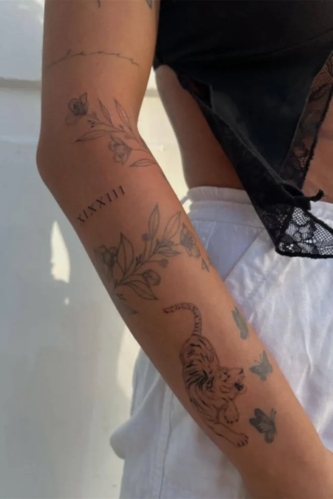 A close-up of a cohesive Y2K sticker sleeve tattoo on a forearm featuring black and grey patchwork designs of flowers, a tiger, and butterflies.