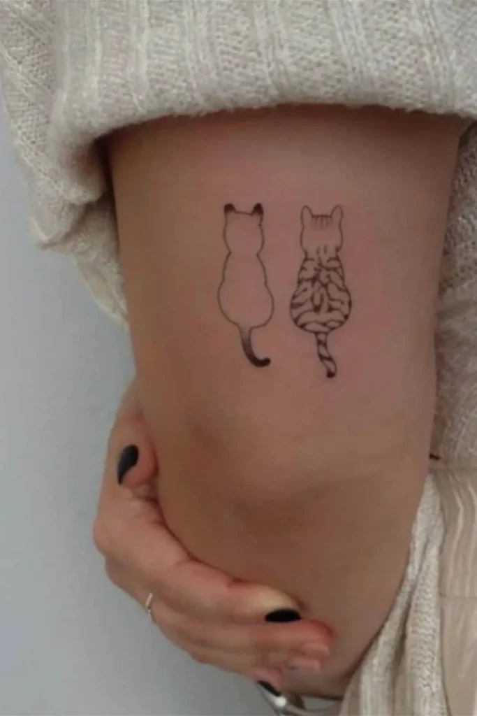 A high-precision Micro Realism Pet Memorial Tattoo on a person's hand featuring two small cat silhouettes viewed from behind, one solid black and one with detailed tabby stripes.