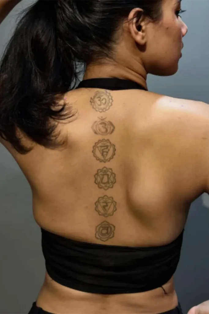 A vertical, fine-line Spine Chakra Sacred Geometry Tattoo with six detailed grey-ink symbols on a woman's upper back.