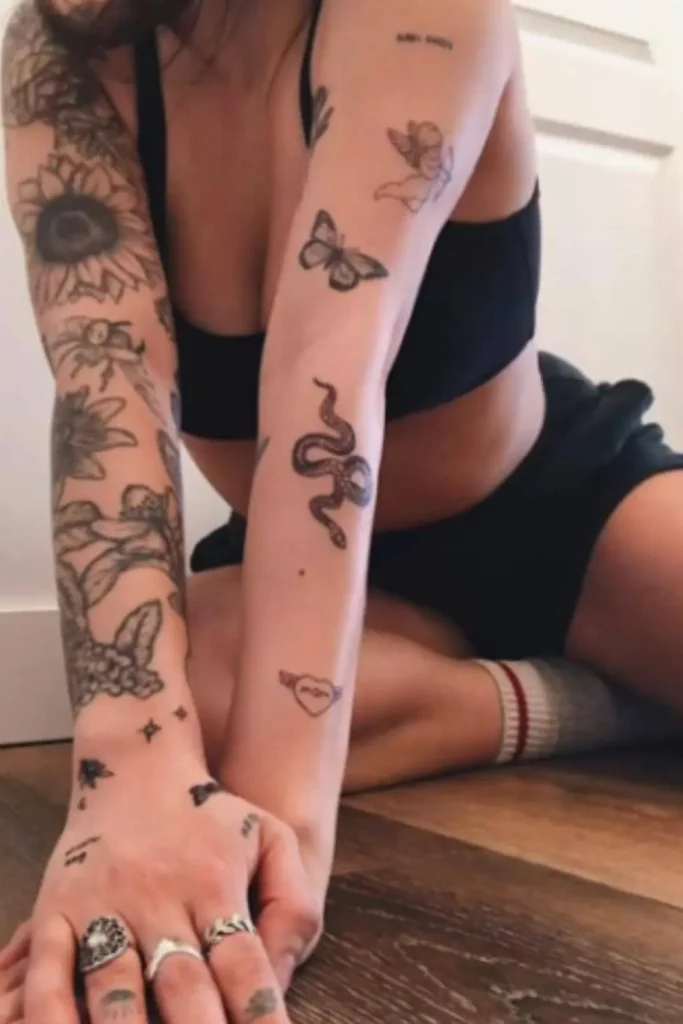 A woman showcasing a growing cohesive Y2K sticker sleeve tattoo with patchwork designs of a snake, butterfly, and cherub on her arms.