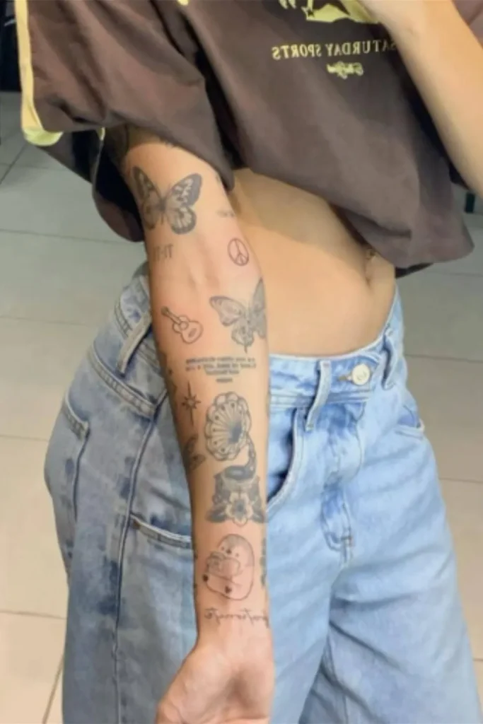 A woman wearing light-wash jeans and a brown t-shirt, showing a forearm covered in a collection of small, fine-line tattoos including butterflies, a guitar, a gramophone, and a peace sign.