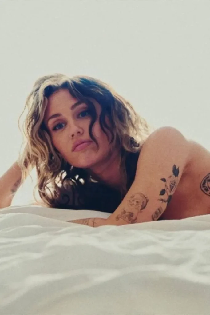 Miley Cyrus is showcasing her cohesive Y2K sticker sleeve tattoo with fine-line patchwork designs on her arm while reclining on white bedding