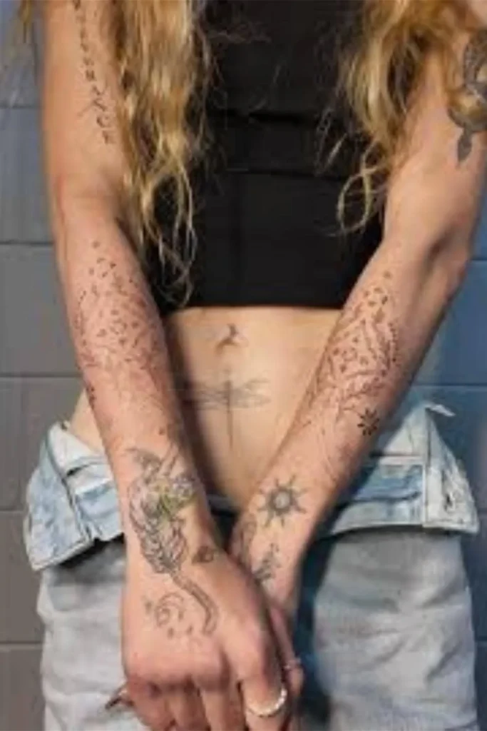 A woman displaying a cohesive Y2K sticker sleeve tattoo with fine-line floral patterns on her forearms and a dragonfly design on her stomach.