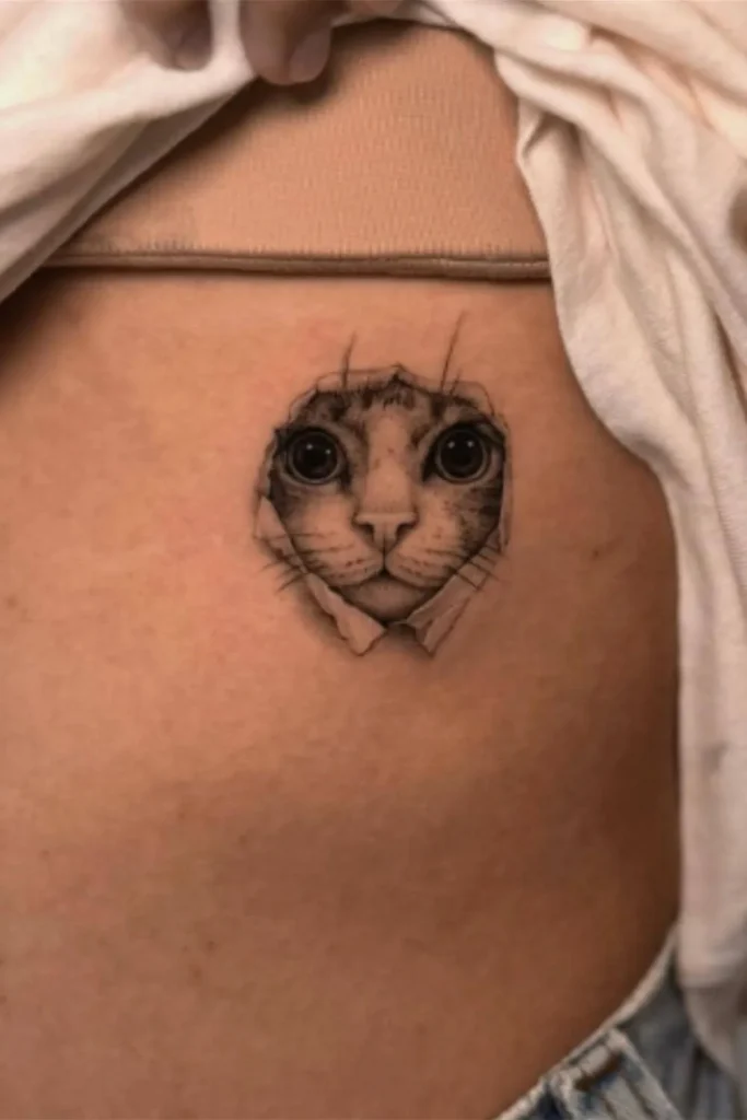 A hyper-realistic Micro Realism Pet Memorial Tattoo on a person's torso featuring a cat's face appearing to peek through a torn skin effect.