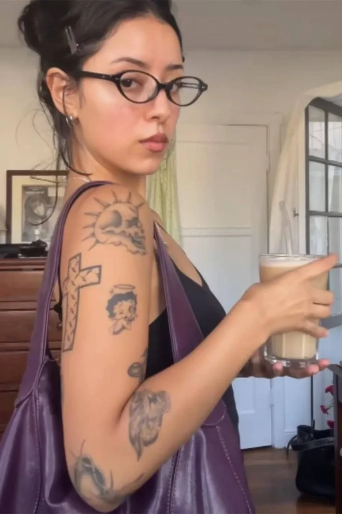 A young woman with glasses and a purple bag holding a drink, showcasing a collection of small tattoos on her arm, including a Micro Realism Pet Memorial Tattoo of a dog's face.