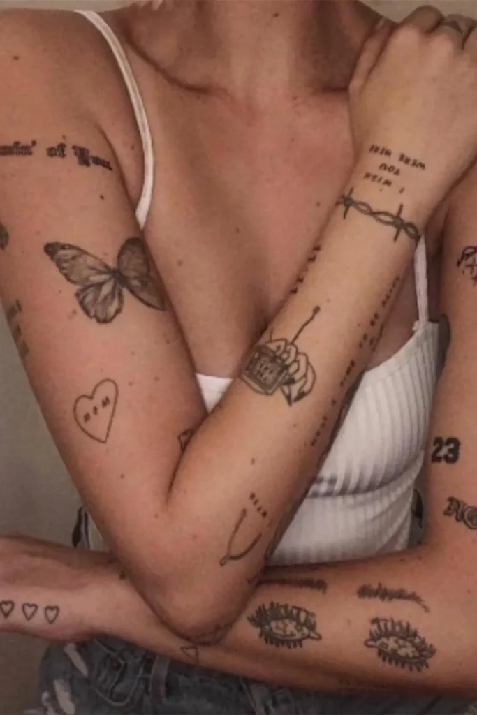 A woman displaying multiple fine-line tattoos on her arms and torso, including small symbols and a butterfly, illustrating how a Micro Realism Pet Memorial Tattoo integrates into a larger collection.