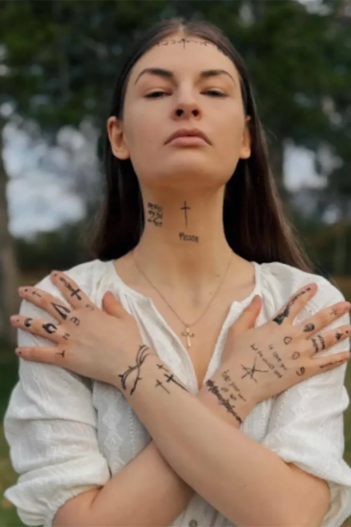 A woman with a cohesive Y2K sticker sleeve tattoo featuring a variety of black-ink patchwork symbols on her arms, hands, and neck.