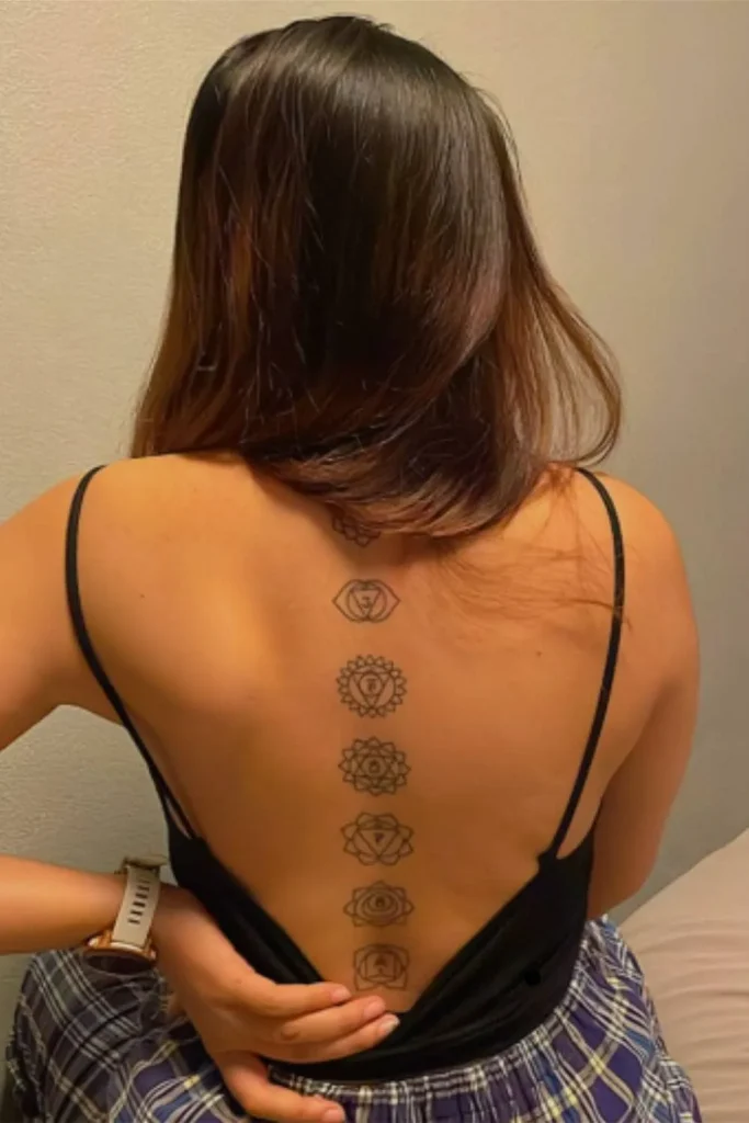 A vertically aligned Spine Chakra Sacred Geometry Tattoo with seven black-ink symbols on a woman's back in a black tank top.
