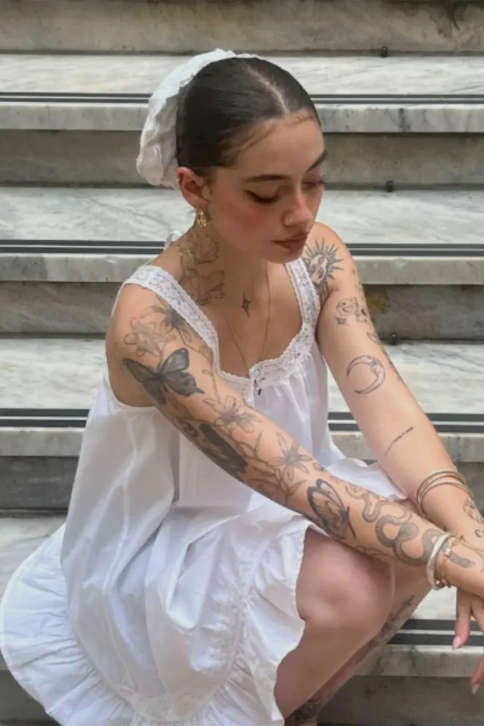 A young woman sitting on stone steps wearing a white dress, showcasing a detailed sleeve of fine-line and botanical tattoos, demonstrating the aesthetic versatility of a Micro Realism Pet Memorial Tattoo.