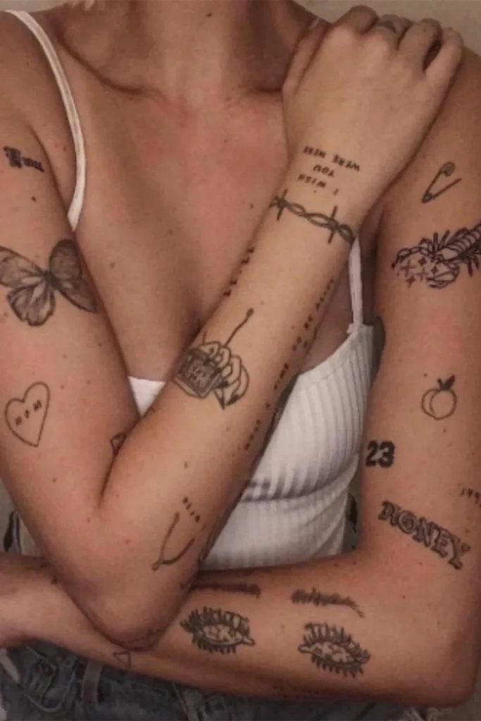 A woman is modeling a cohesive Y2K sticker sleeve tattoo with fine-line patchwork designs of butterflies, hearts, and barbed wire on her arms.