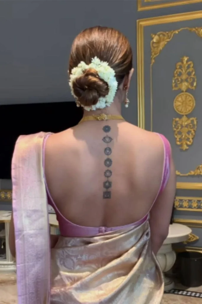 A vertical Spine Chakra Sacred Geometry Tattoo with seven dark grey symbols aligned on a woman's back, worn with a pink and gold traditional outfit.