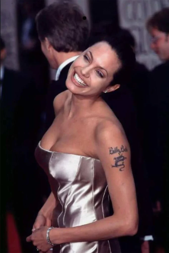 A young Angelina Jolie smiling on the red carpet, showing her early Billy Bob dragon tattoo, illustrating the evolution toward the Angelina Jolie Stay Gold Tattoo Meaning.