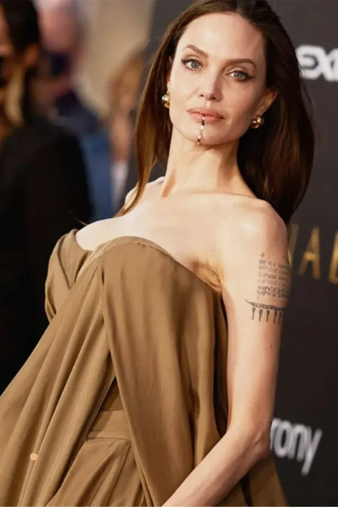 Angelina Jolie wearing a brown strapless gown and a gold chin cuff, with her birth coordinate arm tattoos visible, illustrating the Angelina Jolie Stay Gold Tattoo Meaning.